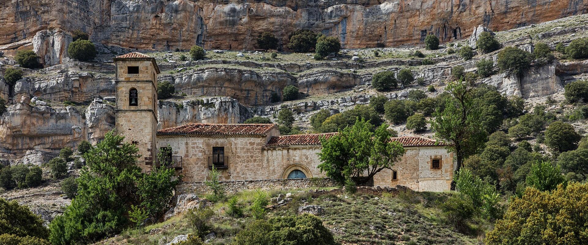 A stone church with a square bell tower and terracotta roof situated on a grassy slope at the base of a large, layered limestone cliff face.