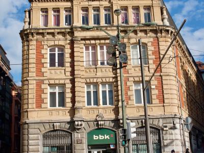 A multi-story historic building with ornate architecture, featuring a large rooftop sign reading "Bilbao Bizkaia Kutxa" and a ground-floor bank entrance with the logo "bbk", located on a city street with pedestrians and traffic infrastructure.