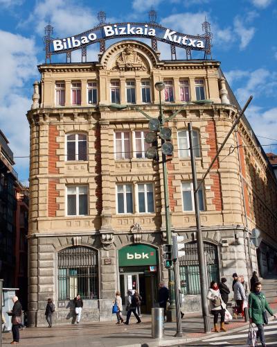 A multi-story historic building with ornate architecture, featuring a large rooftop sign reading "Bilbao Bizkaia Kutxa" and a ground-floor bank entrance with the logo "bbk", located on a city street with pedestrians and traffic infrastructure.