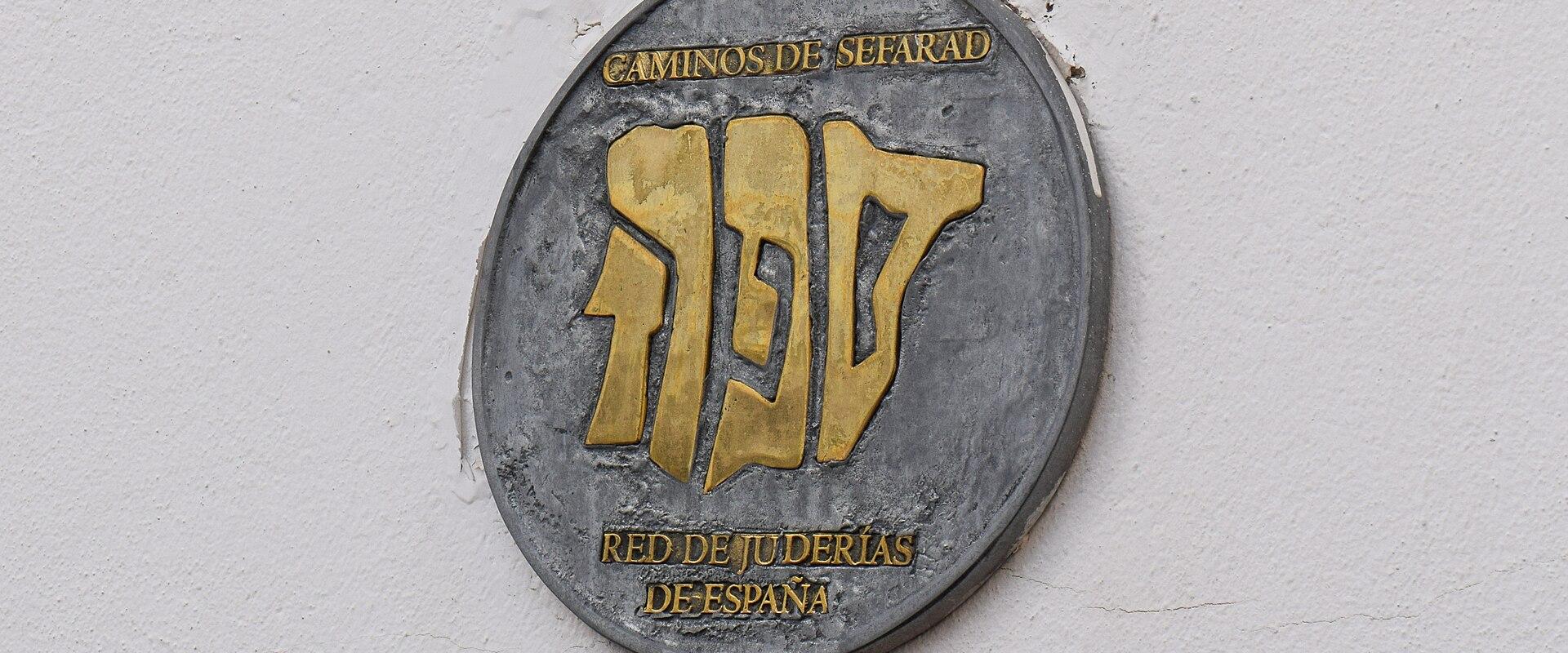 A close-up of a circular metal plaque mounted on a white wall, featuring the Hebrew word "Sefarad" in gold and the text "CAMINOS DE SEFARAD" and "RED DE JUDERÍAS DE ESPAÑA".