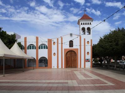 A white church with orange vertical stripes and a bell tower, situated in a paved plaza with white tents on the left and trees on the right under a blue sky.