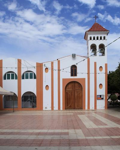 A white church with orange vertical stripes and a bell tower, situated in a paved plaza with white tents on the left and trees on the right under a blue sky.