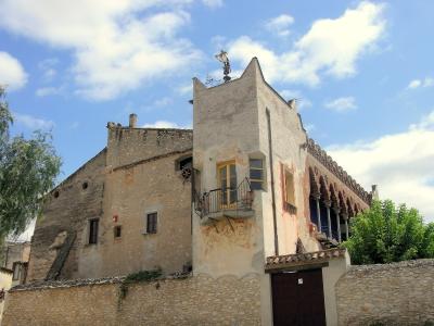 A low-angle view of a historic stone building complex featuring a prominent white tower with a small balcony, a weather vane on the roof, and a long arcade with brick arches to the right, set against a blue sky.