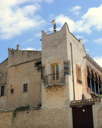 A low-angle view of a historic stone building complex featuring a prominent white tower with a small balcony, a weather vane on the roof, and a long arcade with brick arches to the right, set against a blue sky.