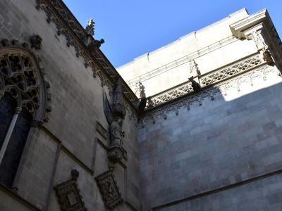 A low-angle view of the exterior stone walls of a Gothic-style building, featuring a large arched window with tracery, a statue of an angel in a niche, and decorative gargoyles along the roofline against a blue sky.