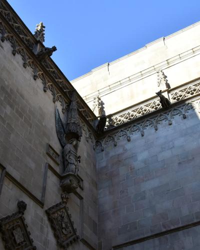 A low-angle view of the exterior stone walls of a Gothic-style building, featuring a large arched window with tracery, a statue of an angel in a niche, and decorative gargoyles along the roofline against a blue sky.