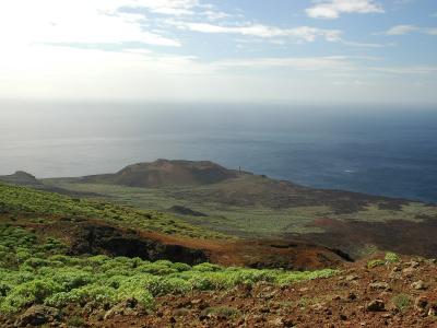 A coastal landscape on a volcanic island showing a white lighthouse standing on a small headland overlooking the ocean, with green scrub and reddish volcanic soil in the foreground.