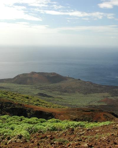 A coastal landscape on a volcanic island showing a white lighthouse standing on a small headland overlooking the ocean, with green scrub and reddish volcanic soil in the foreground.