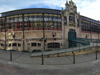 A large, ornate market building with a curved glass facade and stone base, featuring a prominent entrance with a green gate and a sign reading "MERCADO" above it, situated on a paved plaza with bollards and a ramp.