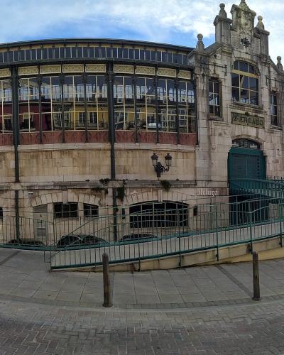A large, ornate market building with a curved glass facade and stone base, featuring a prominent entrance with a green gate and a sign reading "MERCADO" above it, situated on a paved plaza with bollards and a ramp.