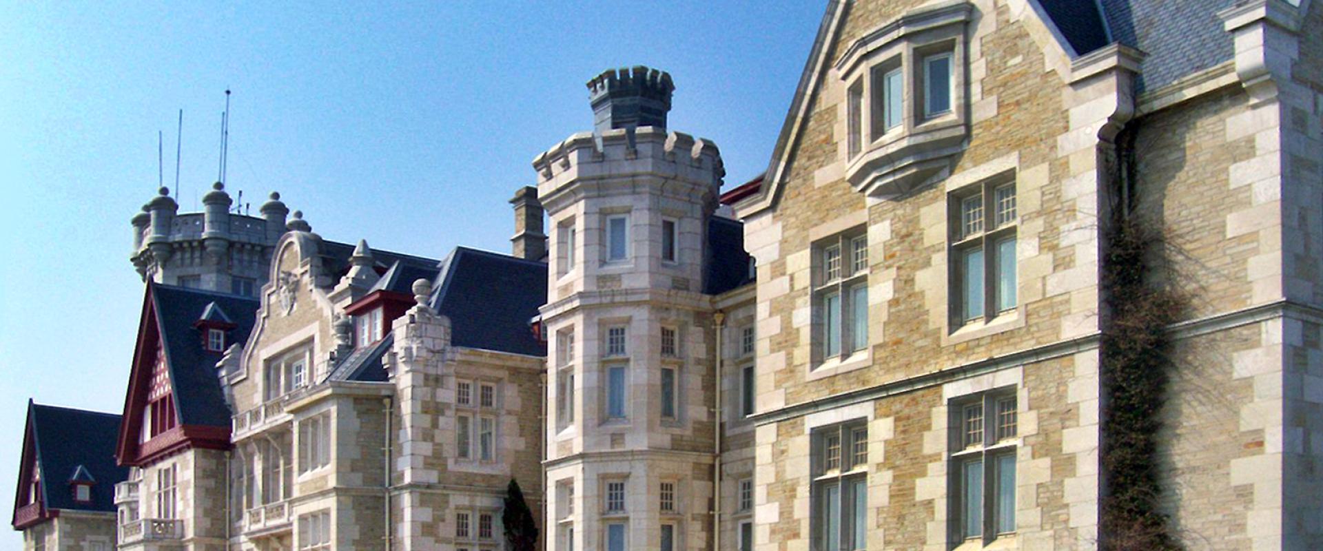 A large, ornate stone building with turrets and a steep roof, set against a clear blue sky, with manicured gardens in the foreground.