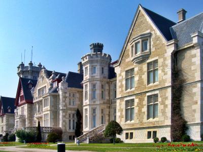 A large, ornate stone building with turrets and a steep roof, set against a clear blue sky, with manicured gardens in the foreground.