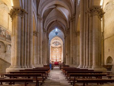The interior nave of a historic stone cathedral featuring Romanesque columns, ribbed vaults, wooden pews, and a brightly colored altarpiece at the far end.