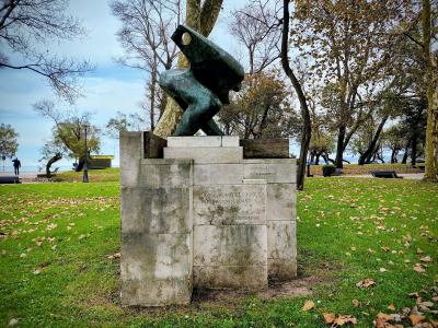 A bronze sculpture of a crouching figure on a stone pedestal in a grassy park, with an inscription reading "AL NOBLE JUEGO DE LOS BOLOS" and "OCTUBRE 1932", set against a backdrop of trees and a glimpse of the sea in the distance.