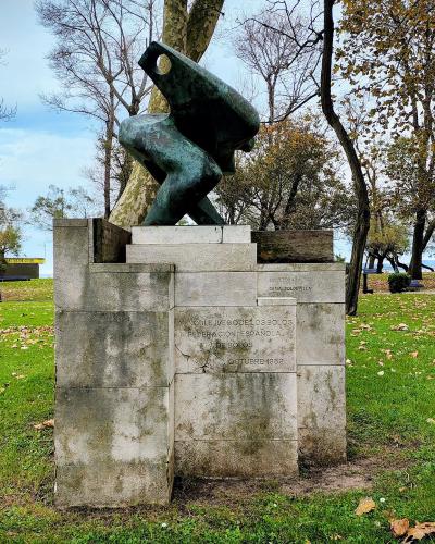 A bronze sculpture of a crouching figure on a stone pedestal in a grassy park, with an inscription reading "AL NOBLE JUEGO DE LOS BOLOS" and "OCTUBRE 1932", set against a backdrop of trees and a glimpse of the sea in the distance.