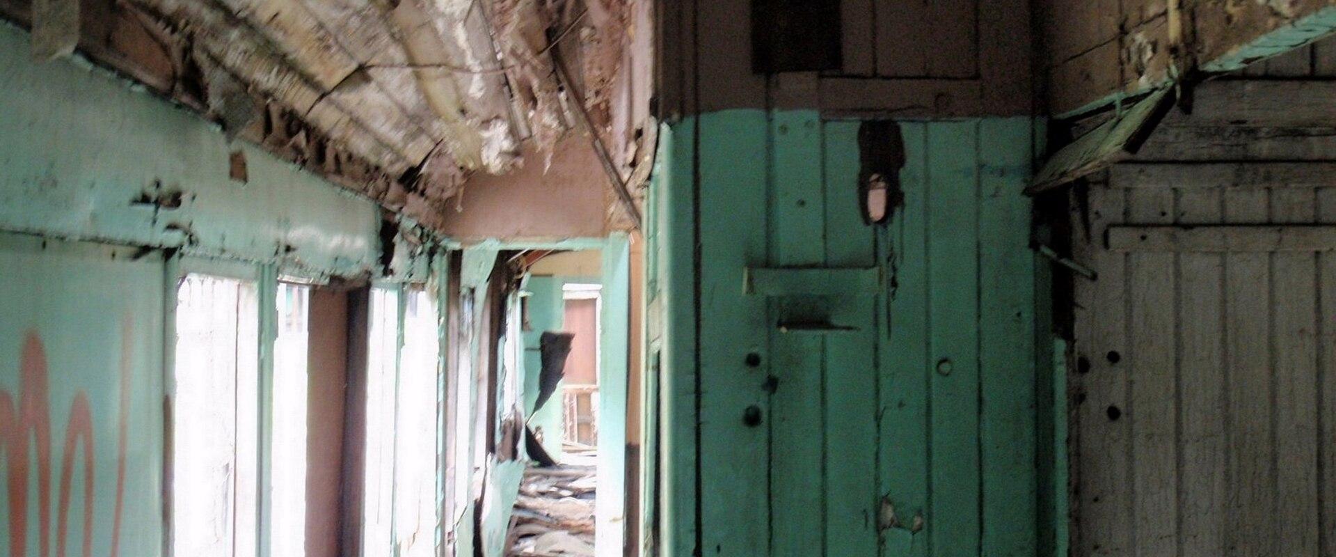 The interior of a dilapidated, abandoned train carriage or railcar, showing peeling teal paint, exposed wooden ceiling beams, and debris on the floor.