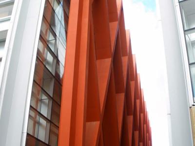 A low-angle architectural shot showing a distinctive facade with large, rust-colored, angular steel beams arranged in a zigzag pattern between white structural columns and glass windows.