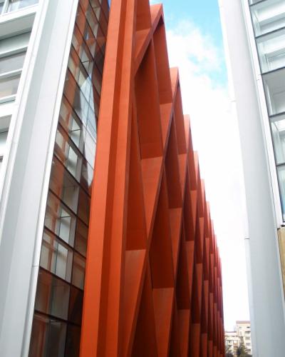 A low-angle architectural shot showing a distinctive facade with large, rust-colored, angular steel beams arranged in a zigzag pattern between white structural columns and glass windows.