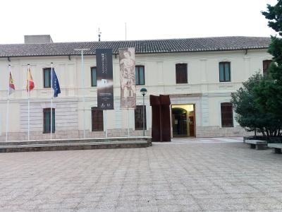 A large, light-colored institutional building with a tiled roof, featuring three flags (Castile-La Mancha, Spain, EU) and two vertical banners advertising an exhibition titled "EL ARTE DE LA GUERRA" (The Art of War) at the "MUSEO DE CIUDAD REAL".