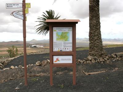 A trailhead signpost and information kiosk on a volcanic landscape, with directional signs pointing to "Ermita de Las Nieves" and "Peñas del Chache".