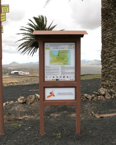A trailhead signpost and information kiosk on a volcanic landscape, with directional signs pointing to "Ermita de Las Nieves" and "Peñas del Chache".