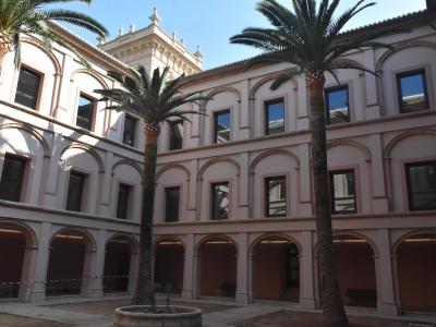 A sunlit interior courtyard of a classical-style building featuring two-story arcades with pinkish stucco walls, arched colonnades on the ground floor, and tall palm trees planted in a central stone basin.