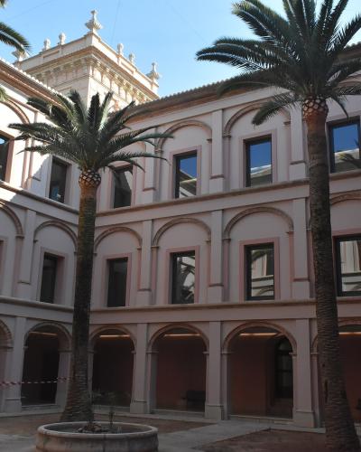 A sunlit interior courtyard of a classical-style building featuring two-story arcades with pinkish stucco walls, arched colonnades on the ground floor, and tall palm trees planted in a central stone basin.