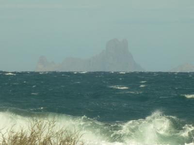 A rocky island formation rising from a choppy sea, viewed from a shoreline with dry grass in the foreground under an overcast sky.