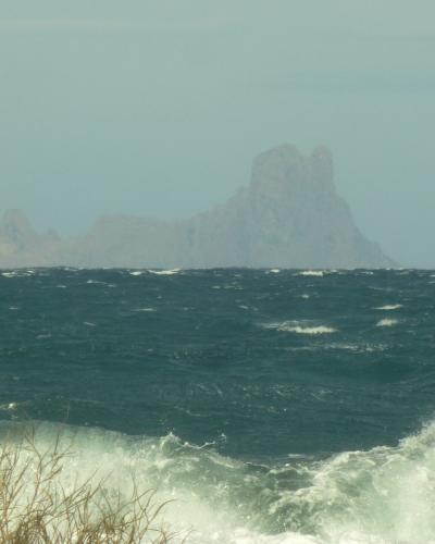 A rocky island formation rising from a choppy sea, viewed from a shoreline with dry grass in the foreground under an overcast sky.