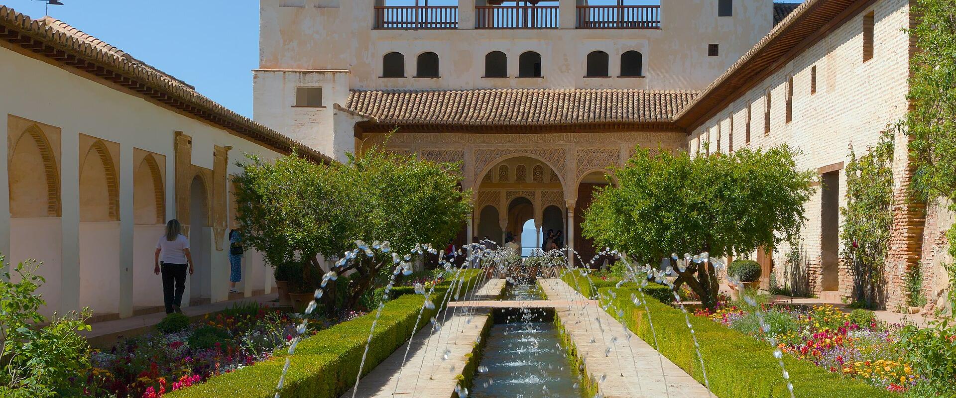 A long rectangular water channel lined with fountains and manicured hedges, flanked by walkways and buildings with Moorish arches and tiled roofs under a clear blue sky.