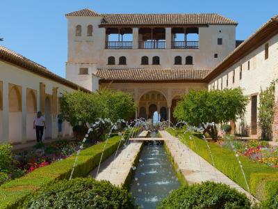 A long rectangular water channel lined with fountains and manicured hedges, flanked by walkways and buildings with Moorish arches and tiled roofs under a clear blue sky.
