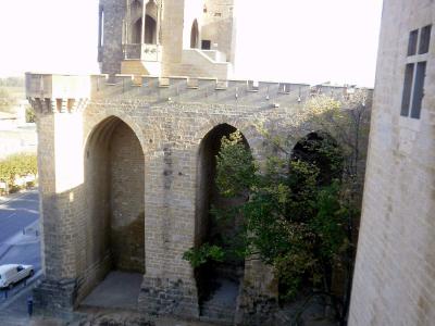 A high-angle view of the exterior stone walls, arched gateways, and a crenellated tower of the Royal Palace of Olite, with a street and white van visible below.