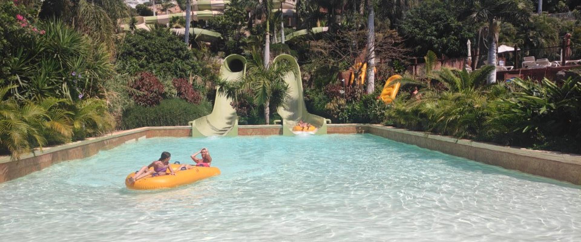A water park scene featuring a lazy river with people on an inflatable raft, two green tube slides, and a large brown temple-like structure in the background surrounded by palm trees.