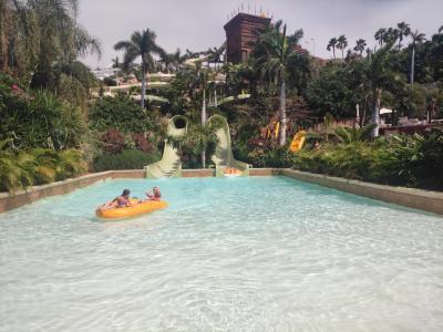 A water park scene featuring a lazy river with people on an inflatable raft, two green tube slides, and a large brown temple-like structure in the background surrounded by palm trees.