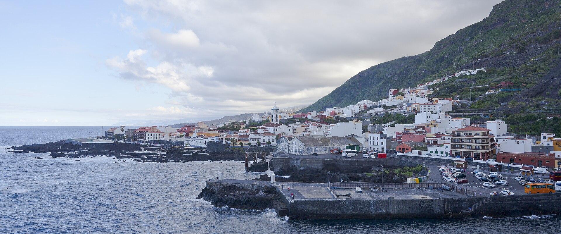 A coastal town with white buildings and a prominent church tower situated along a rocky shoreline with a stone pier extending into the ocean, backed by a steep green hillside.