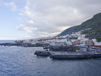 A coastal town with white buildings and a prominent church tower situated along a rocky shoreline with a stone pier extending into the ocean, backed by a steep green hillside.