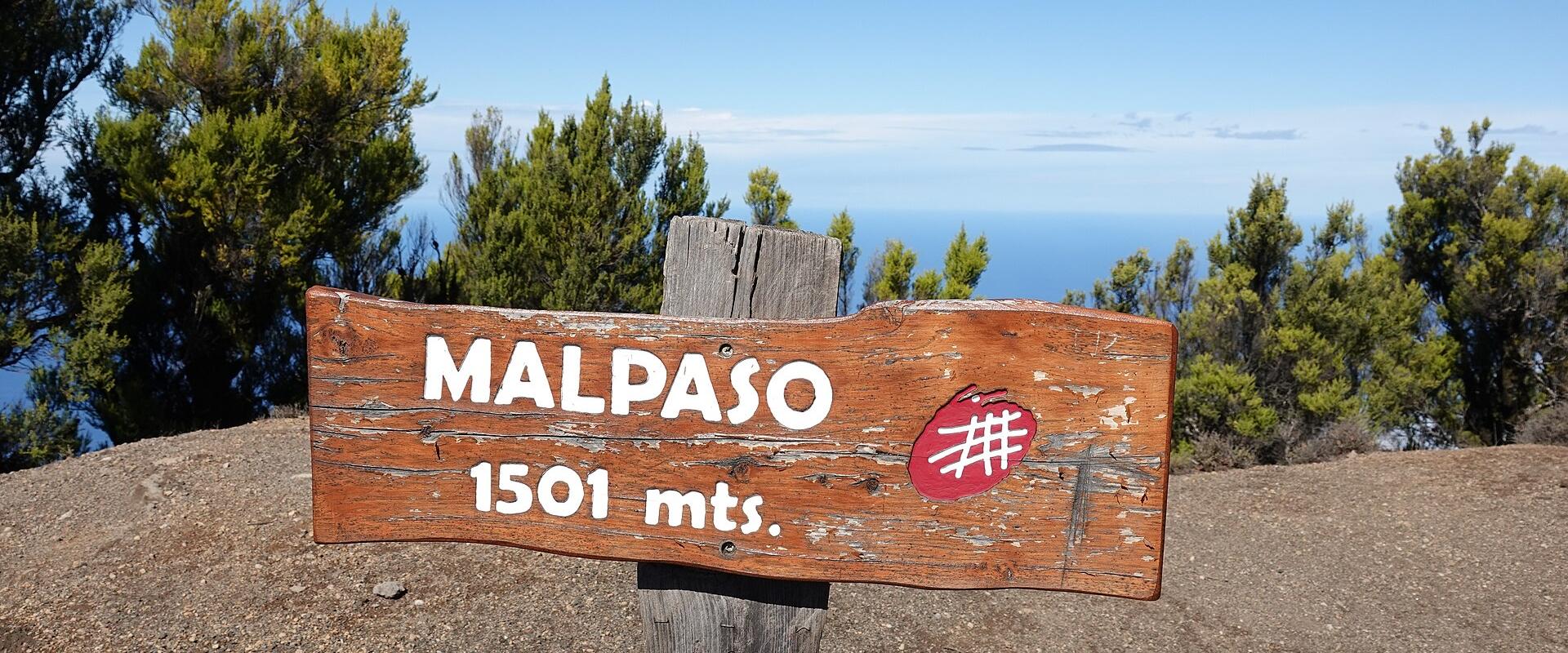 A weathered wooden trail sign reading "MALPASO 1501 mts." with a red hiking trail marker, set against a backdrop of green trees and a distant ocean horizon.