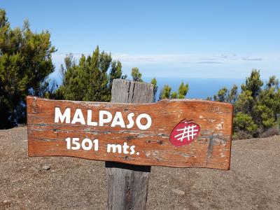 A weathered wooden trail sign reading "MALPASO 1501 mts." with a red hiking trail marker, set against a backdrop of green trees and a distant ocean horizon.