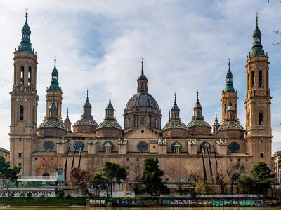 A wide-angle photograph of a large, ornate Baroque cathedral with two prominent bell towers, multiple domes with colorful tilework, and a central dome, situated next to a river with graffiti-covered embankments.