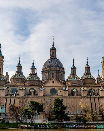 A wide-angle photograph of a large, ornate Baroque cathedral with two prominent bell towers, multiple domes with colorful tilework, and a central dome, situated next to a river with graffiti-covered embankments.