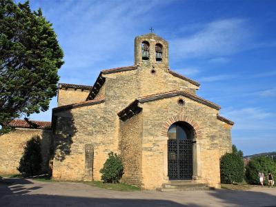 A stone church with a distinctive bell tower featuring two bells, arched entrance with iron gate, and surrounding greenery under a blue sky.