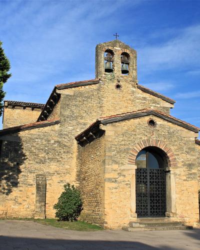 A stone church with a distinctive bell tower featuring two bells, arched entrance with iron gate, and surrounding greenery under a blue sky.
