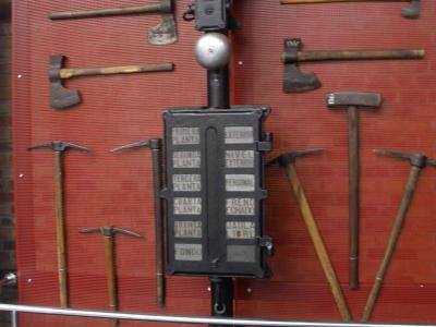 A vintage mining signal lamp and control box mounted on a pole, surrounded by various axes and pickaxes displayed on a red mesh wall.