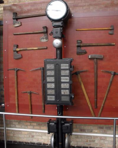 A vintage mining signal lamp and control box mounted on a pole, surrounded by various axes and pickaxes displayed on a red mesh wall.