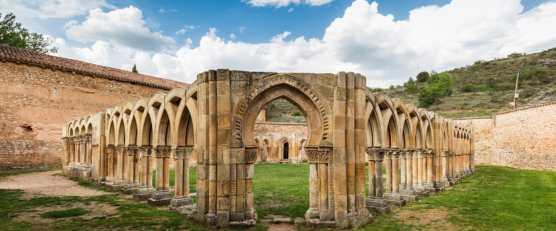 A panoramic view of the ruins of a Romanesque cloister featuring a distinctive central horseshoe arch and rows of columns with carved capitals, set within a grassy courtyard under a blue sky.