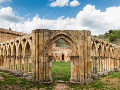 A panoramic view of the ruins of a Romanesque cloister featuring a distinctive central horseshoe arch and rows of columns with carved capitals, set within a grassy courtyard under a blue sky.