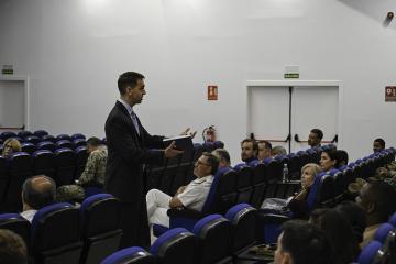A man in a suit is giving a lecture to an audience seated in blue chairs inside a plain white room with "SALIDA" signs above doors.