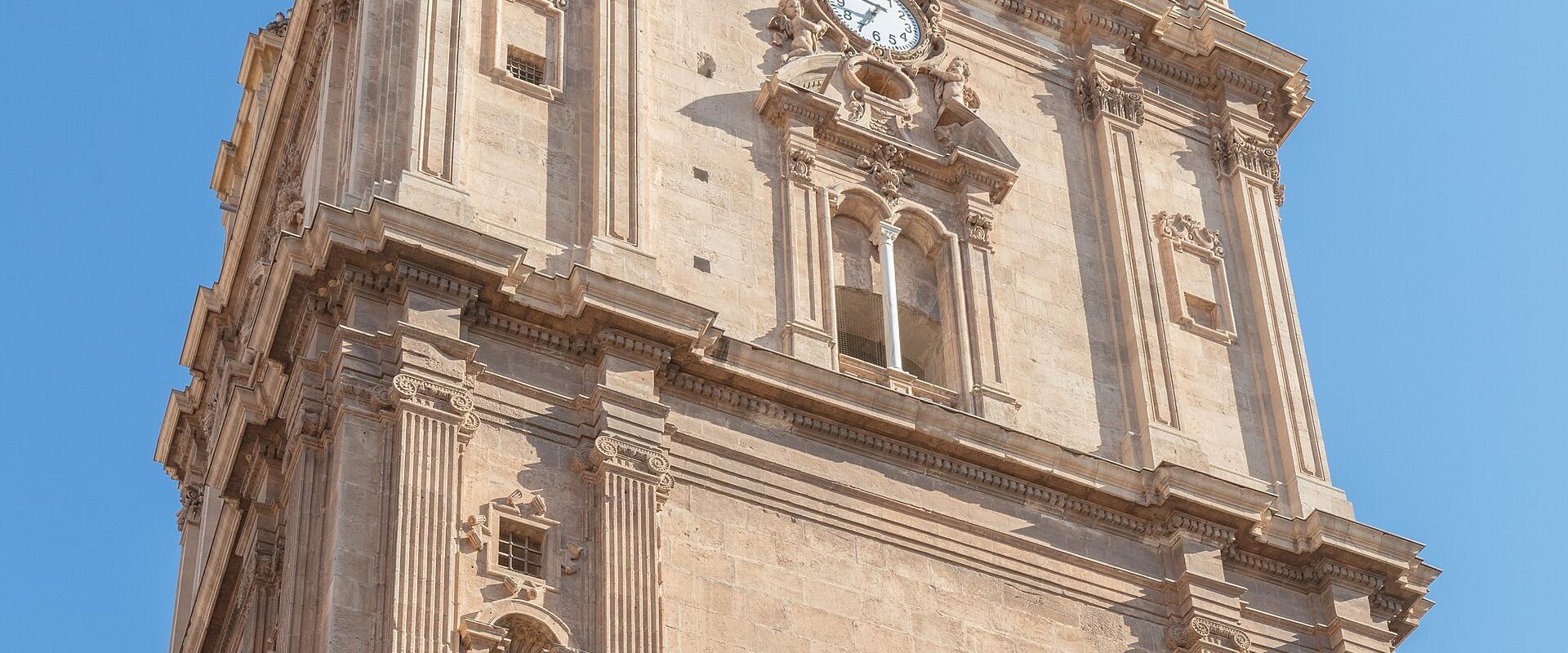 A low-angle shot of an ornate, multi-tiered stone bell tower featuring a clock face, arched belfry openings, and Baroque-style sculptural details against a clear blue sky.