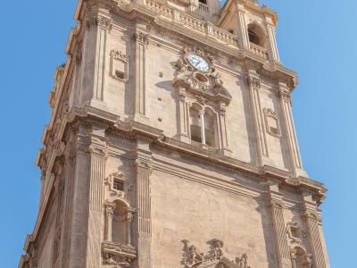 A low-angle shot of an ornate, multi-tiered stone bell tower featuring a clock face, arched belfry openings, and Baroque-style sculptural details against a clear blue sky.