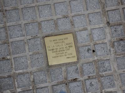 A close-up of a brass commemorative plaque embedded in a paved sidewalk, reading "En esta casa vivió MANUEL DE LA CALLE CAMAS... RMHU-2020".